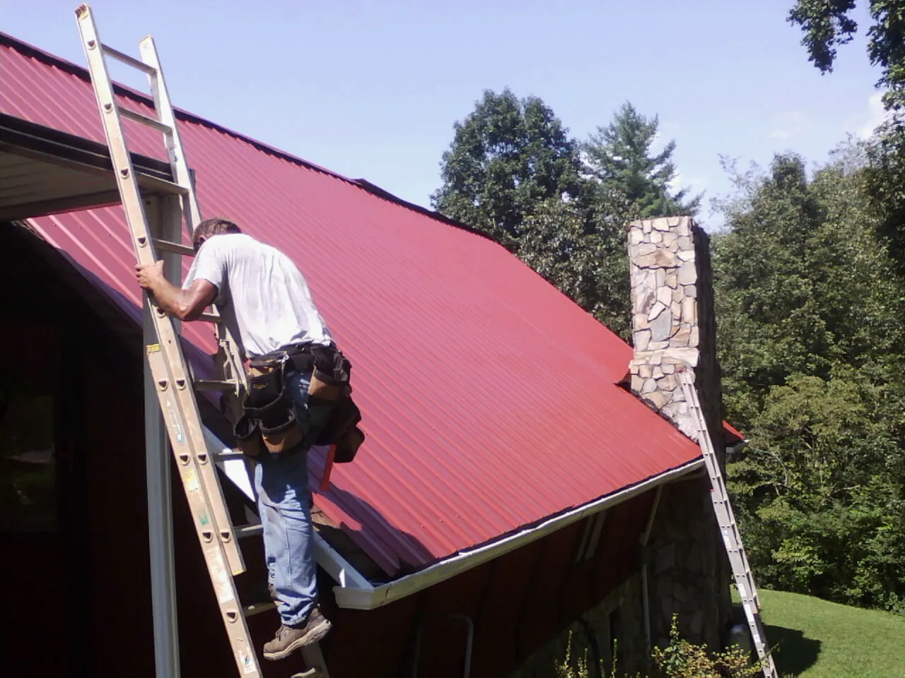 Roofer on a ladder beside a red metal roof and stone chimney — Soffit & Fascia Repair by Roofing Brothers in Unityville
